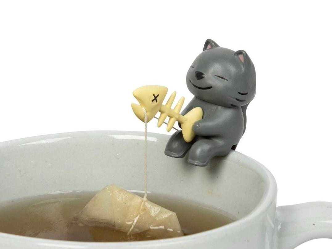 Small grey ceramic cat figurine perched on a mug rim holding a fish-skeleton rod that dangles a tea bag