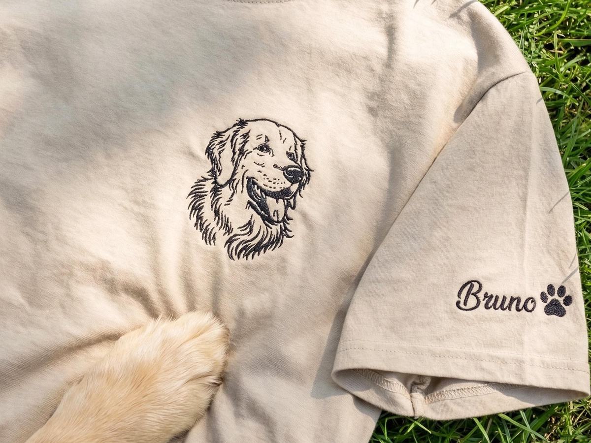 Tan t-shirt with an embroidered line drawing of a golden retriever on the chest and a name with a paw print on the sleeve