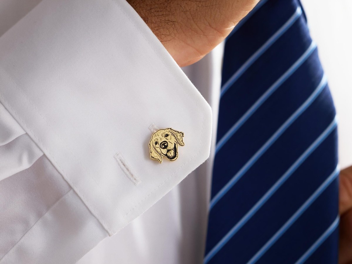Gold-finish cufflink engraved with a smiling dog's face worn on a white shirt cuff beside a blue striped tie