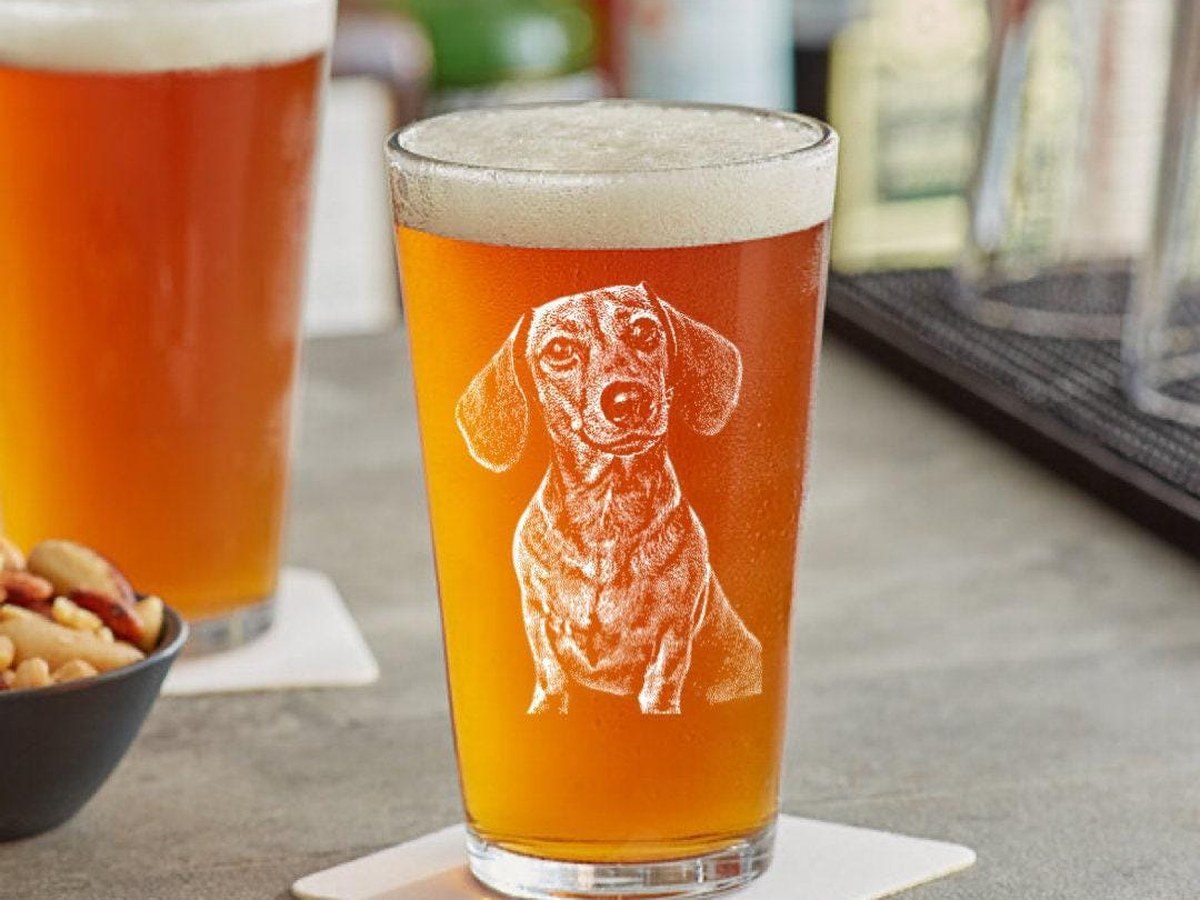 Pint glass of amber beer with a dachshund's face engraved in white on the side, on a bar coaster