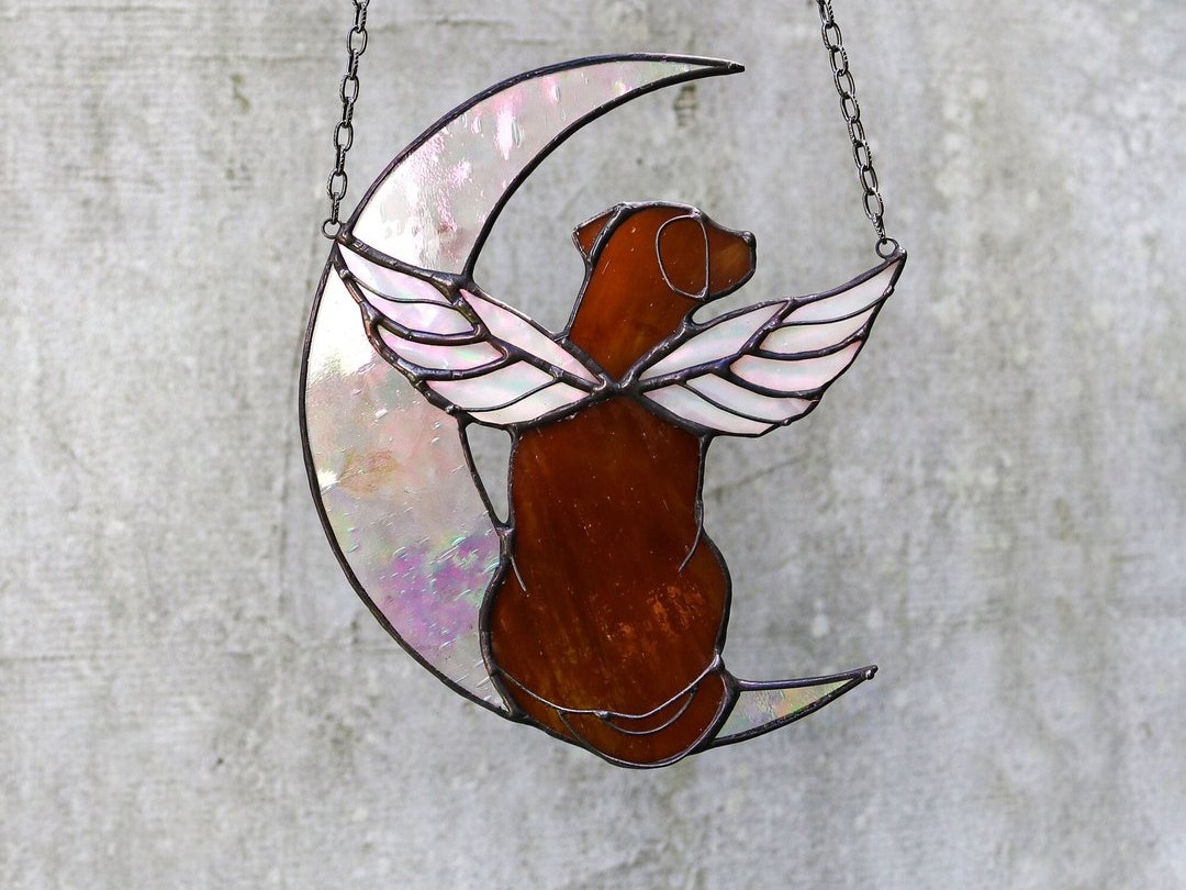Stained glass suncatcher of a winged dog sitting on a crescent moon, hung in a window