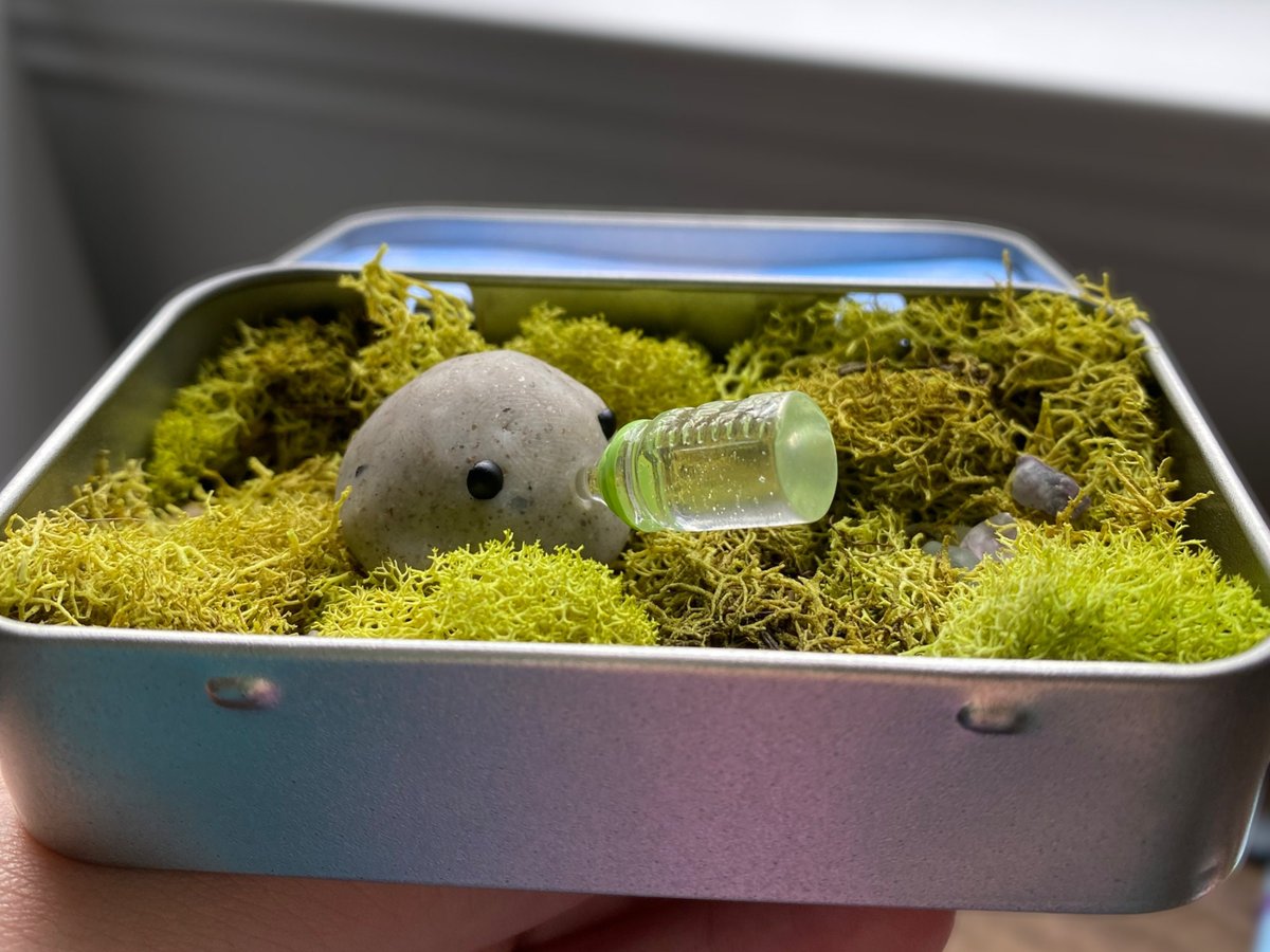 Grey polymer clay pet rock with a tiny green baby bottle, sitting on moss in a metal mint tin