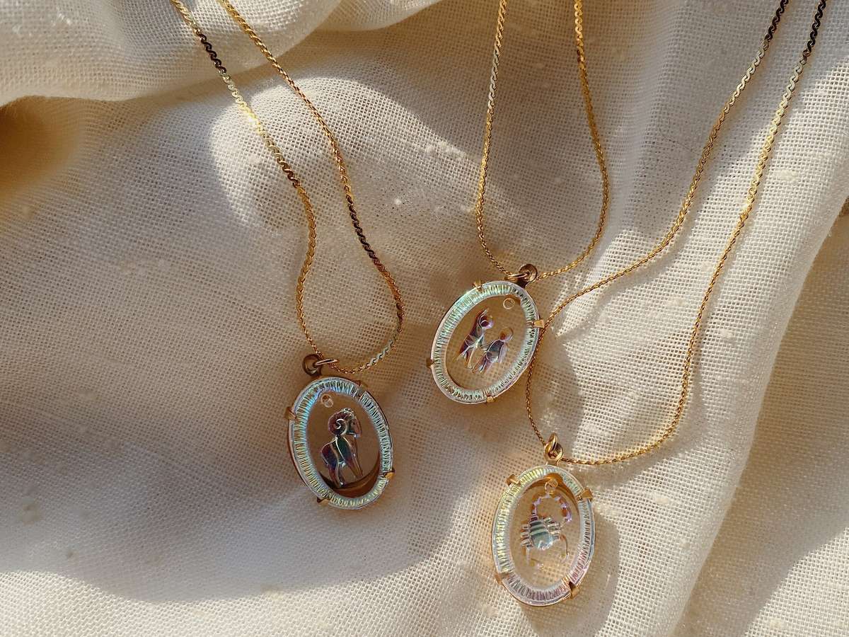 Three oval iridescent glass zodiac pendants (Aries, Gemini, Scorpio) on gold-tone chains on cream linen