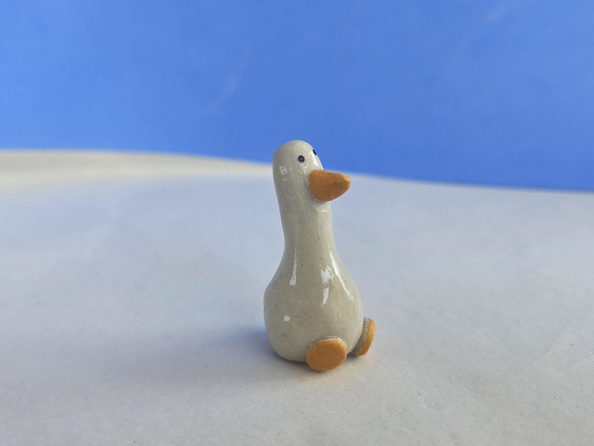 Tiny white stoneware goose figurine with an orange beak and feet on a pale surface